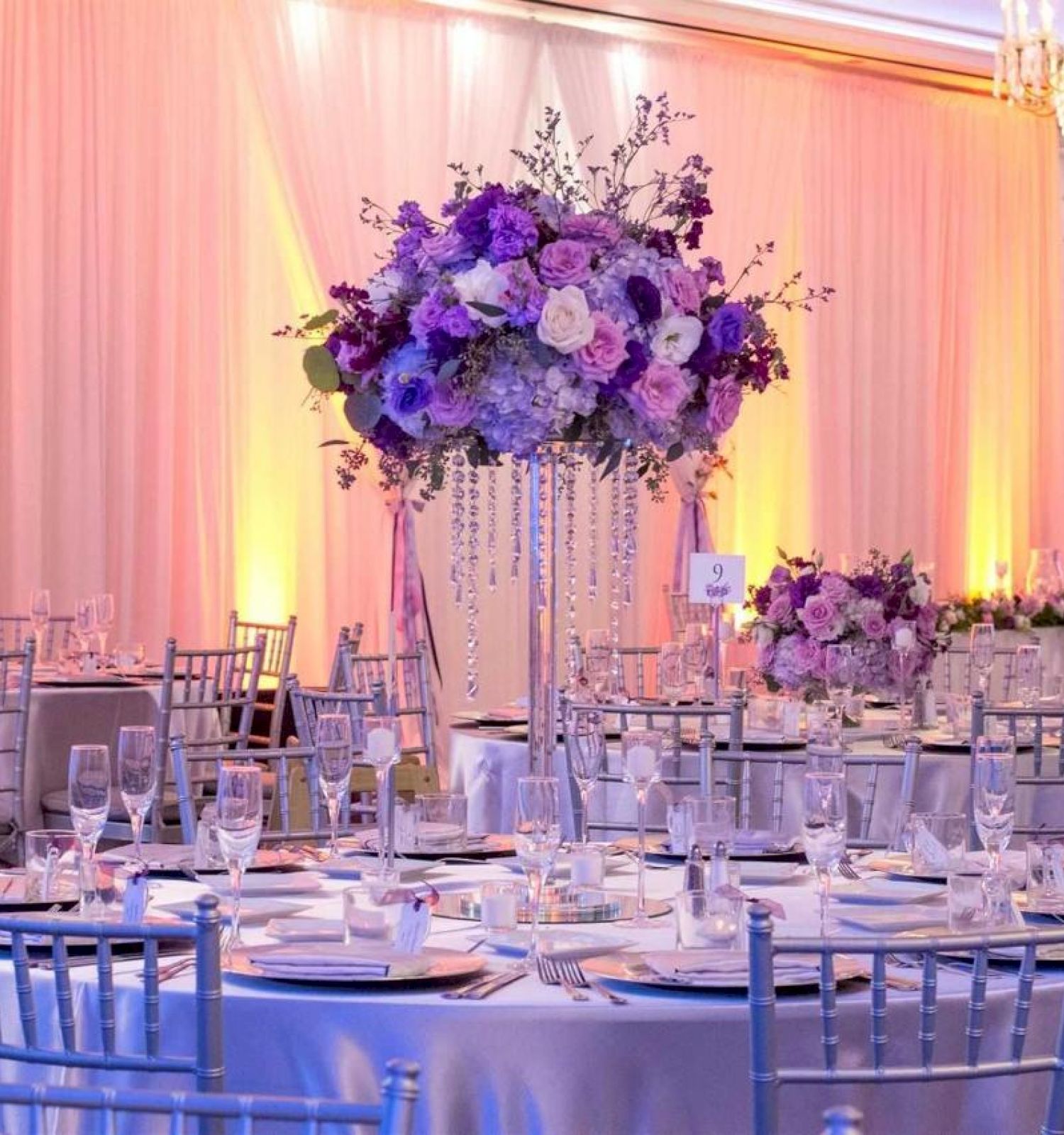 The image shows an elegantly decorated event space with round tables, silver chairs, and purple and white floral centerpieces.