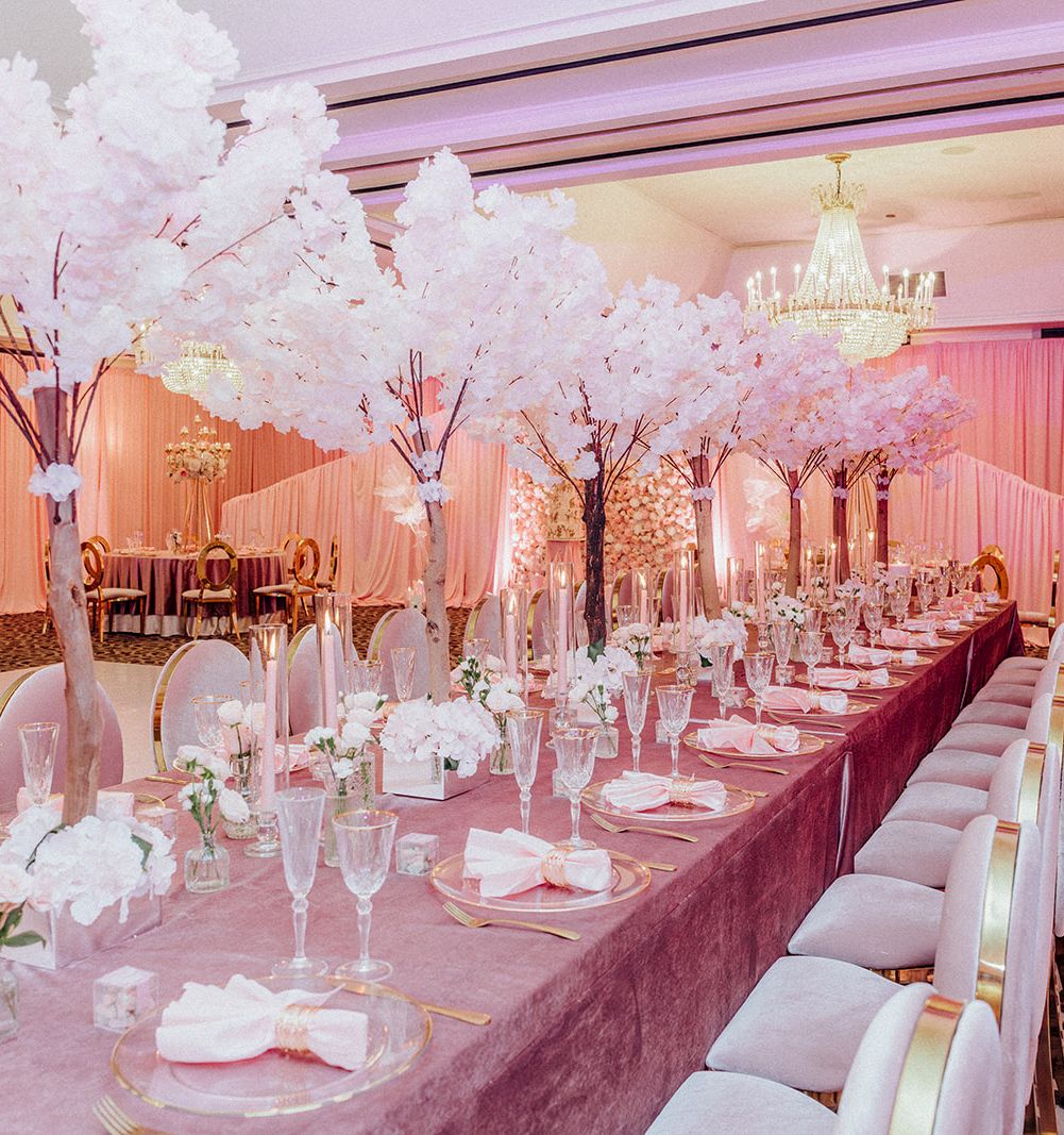 A pink-and-white banquet setup for a wedding or event, with long tables, chandeliers, floral trees, and place settings.