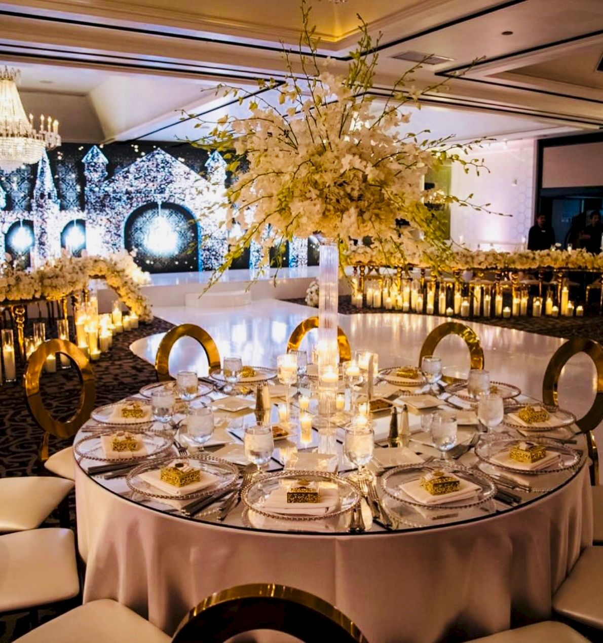Elegant banquet setup with round table candles, gold chair accents, and golden floral arrangements under chandeliers.