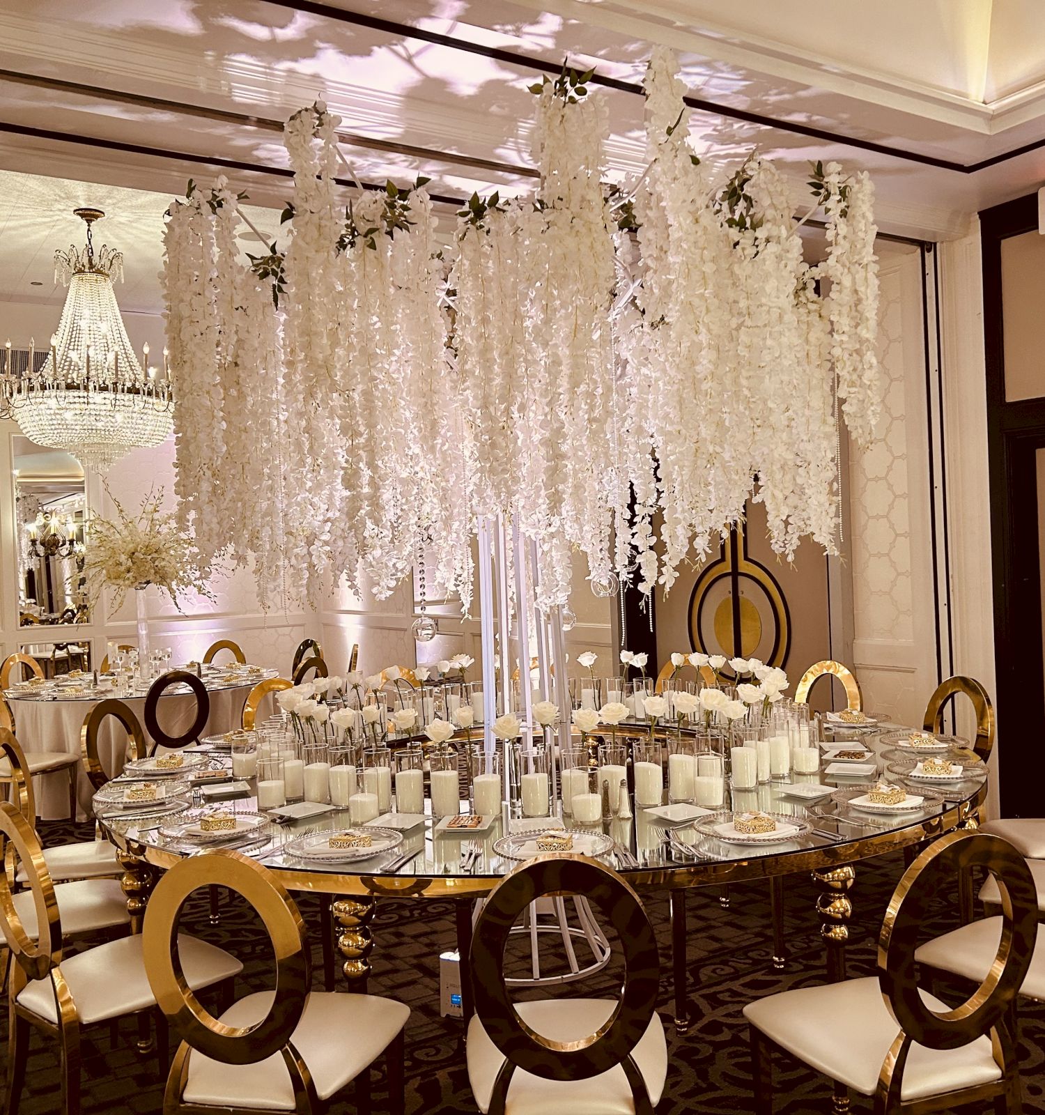 A lavish banquet setup with gold chairs, candlelit tables, and cascading white floral drapes with a chandelier overhead.