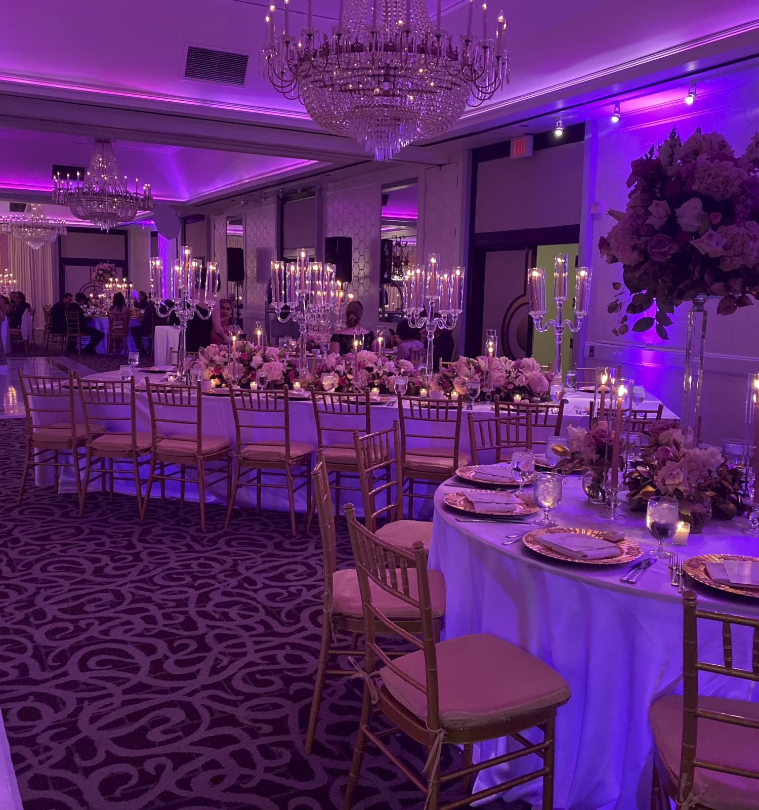 Elegant banquet hall set for a formal event, featuring chandeliers, purple lighting, candlelit tables, and abundant white floral centerpieces.