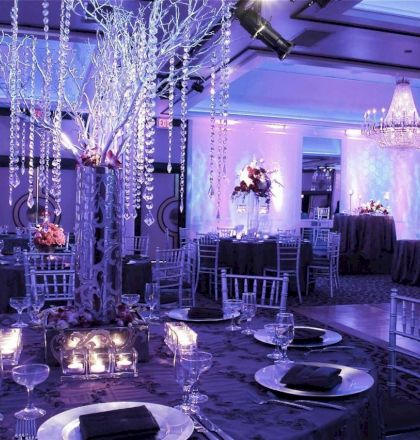 A beautifully decorated event hall with round tables set for dinner, featuring chandeliers and blue ambient lighting, creating an elegant atmosphere.