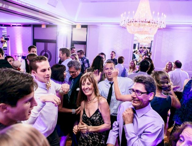 A group of people is dancing and socializing at an indoor event or party under a chandelier with purple lighting.