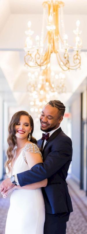 A couple stands in a hallway with chandeliers, the man in a suit embraces the woman in a white dress, both smiling.