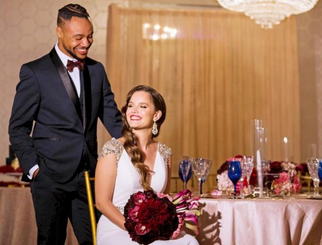 A man in a suit stands beside a seated woman in a white dress holding a bouquet; they are in an elegantly decorated room with a chandelier.