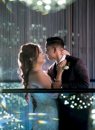 A couple is standing on a balcony, embracing and gazing into each other's eyes amidst a backdrop of decorative lights and reflections.