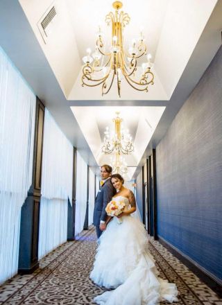 A newlywed couple in wedding attire is posing in an elegant hallway with ornate chandeliers and patterned carpeting, looking happy and in love.