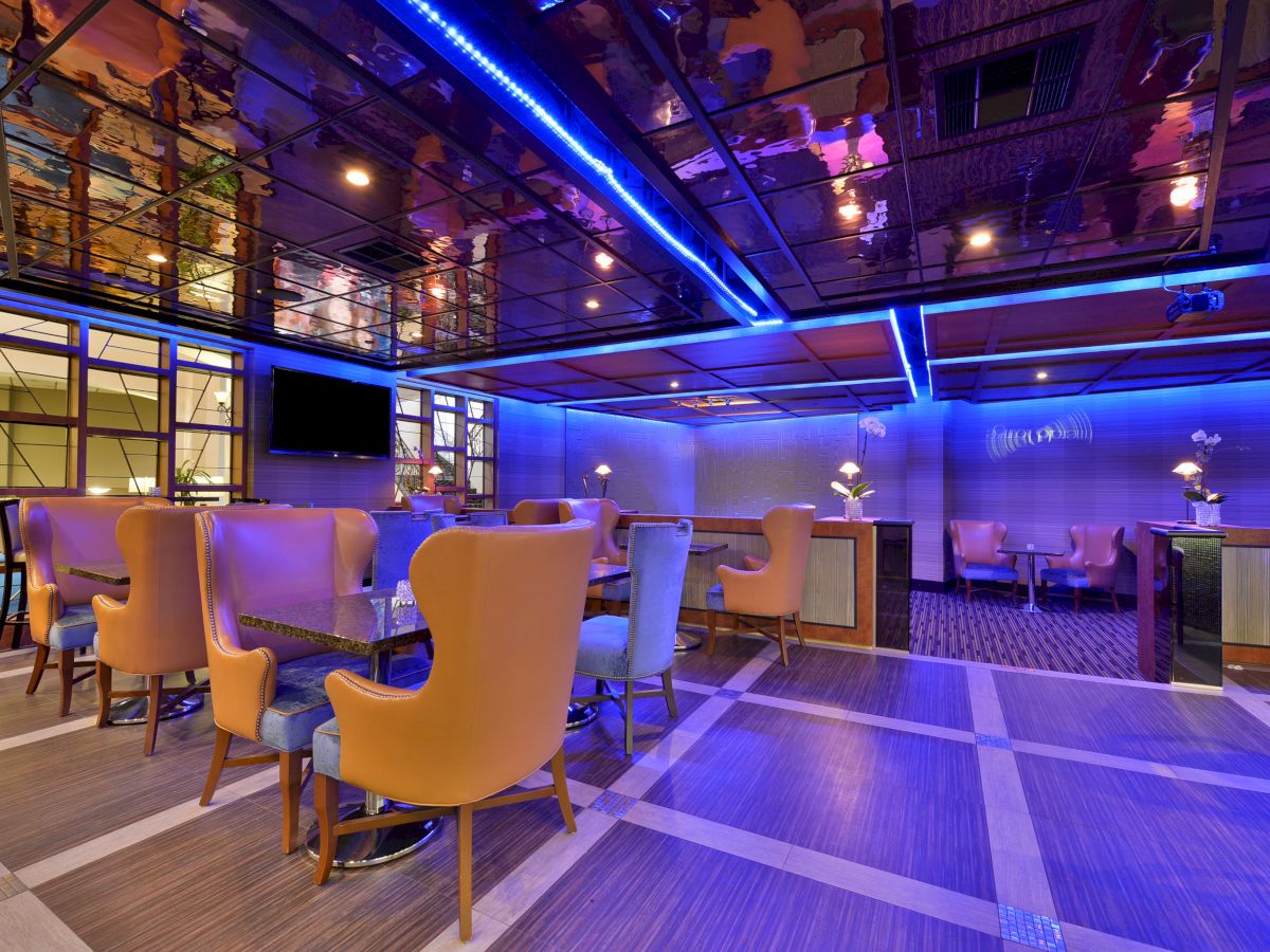 A modern lounge with vibrant lighting, upholstered chairs, tables, a TV, and stylish decor with purple and blue tones, seating areas are visible.