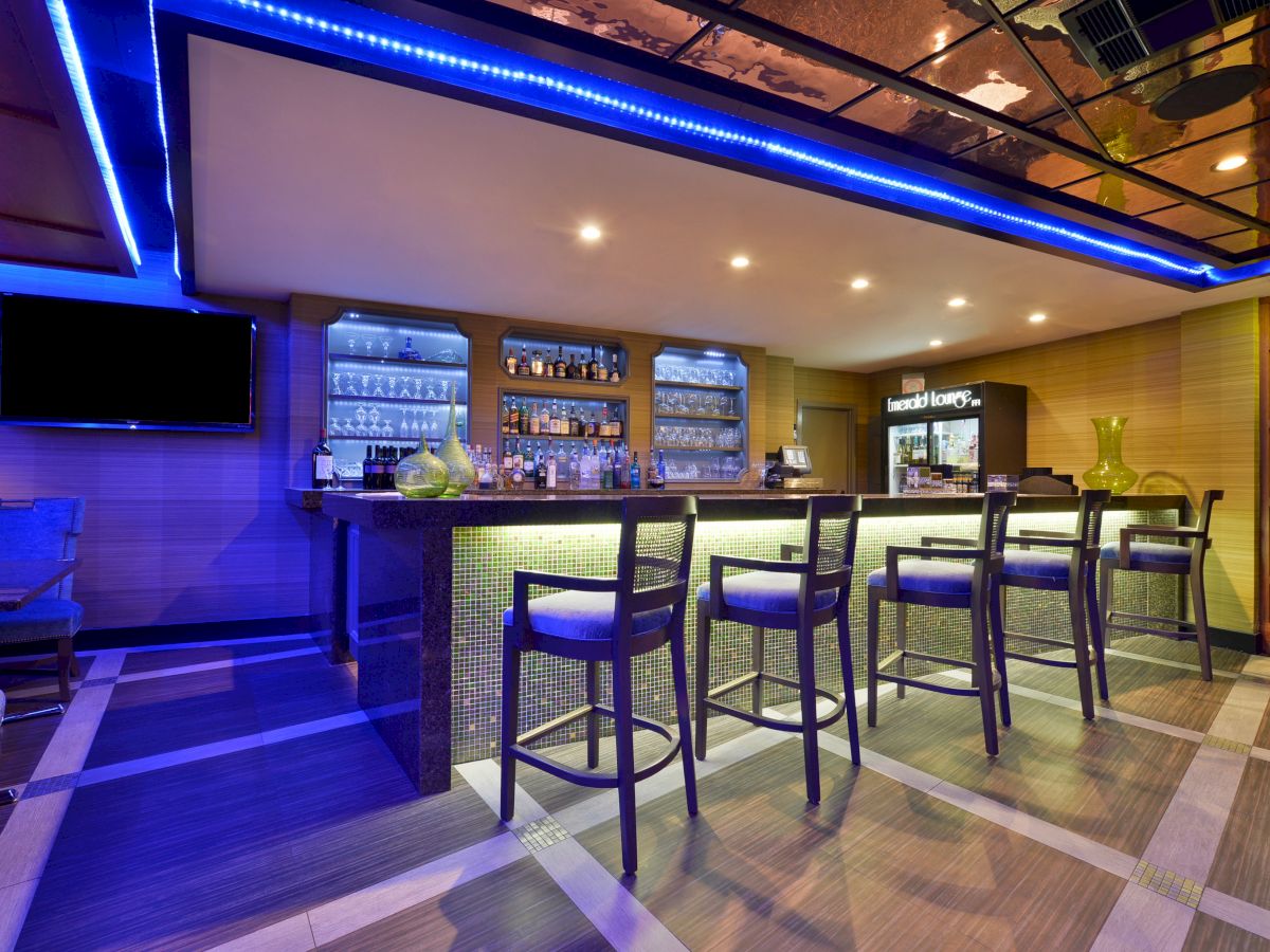 This image shows a modern bar with high chairs, a counter, a shelf stocked with bottles, and blue ambient lighting.