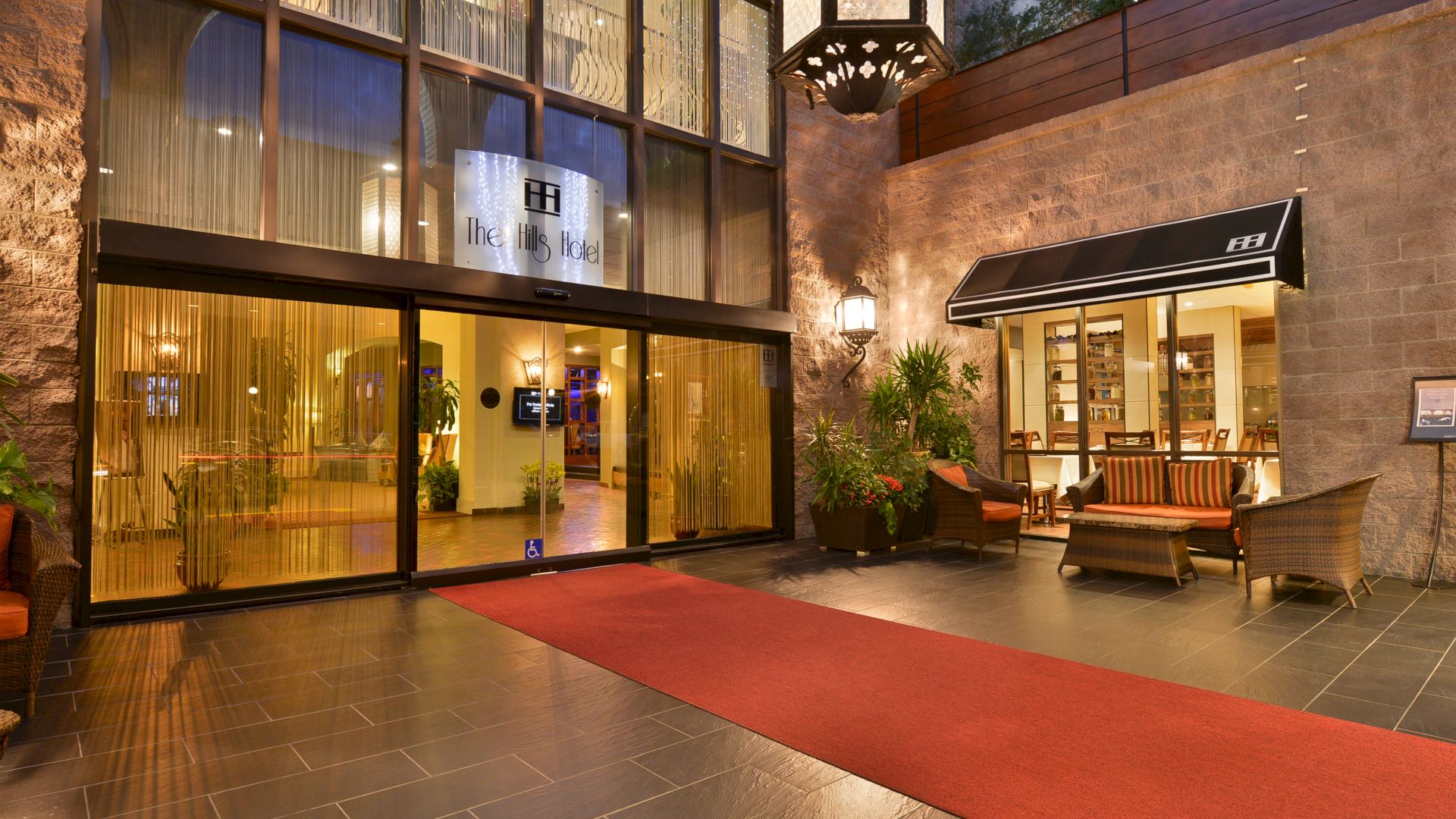 A well-lit hotel entrance with glass doors, a red carpet, outdoor seating area with wicker furniture, plants, and a decorative wall lamp.