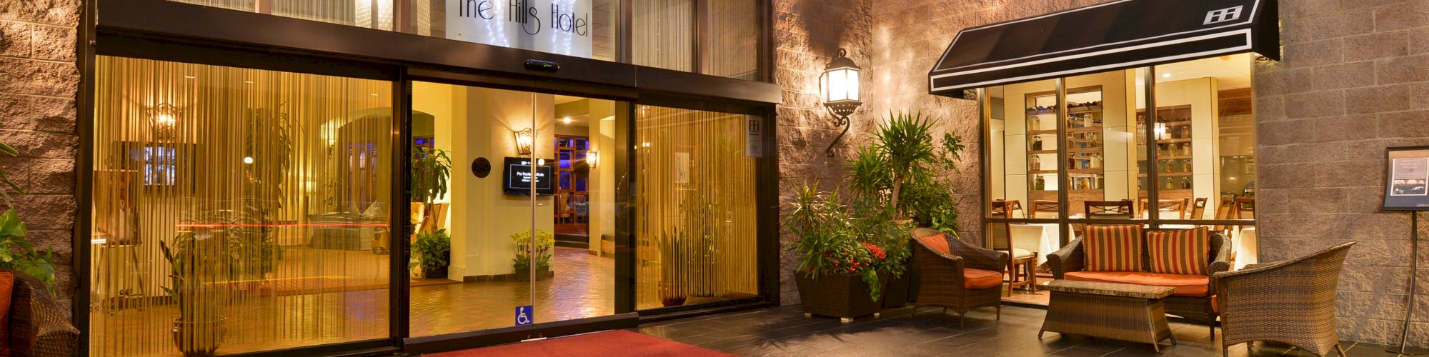 A well-lit hotel entrance with glass doors, a red carpet, outdoor seating area with wicker furniture, plants, and a decorative wall lamp.