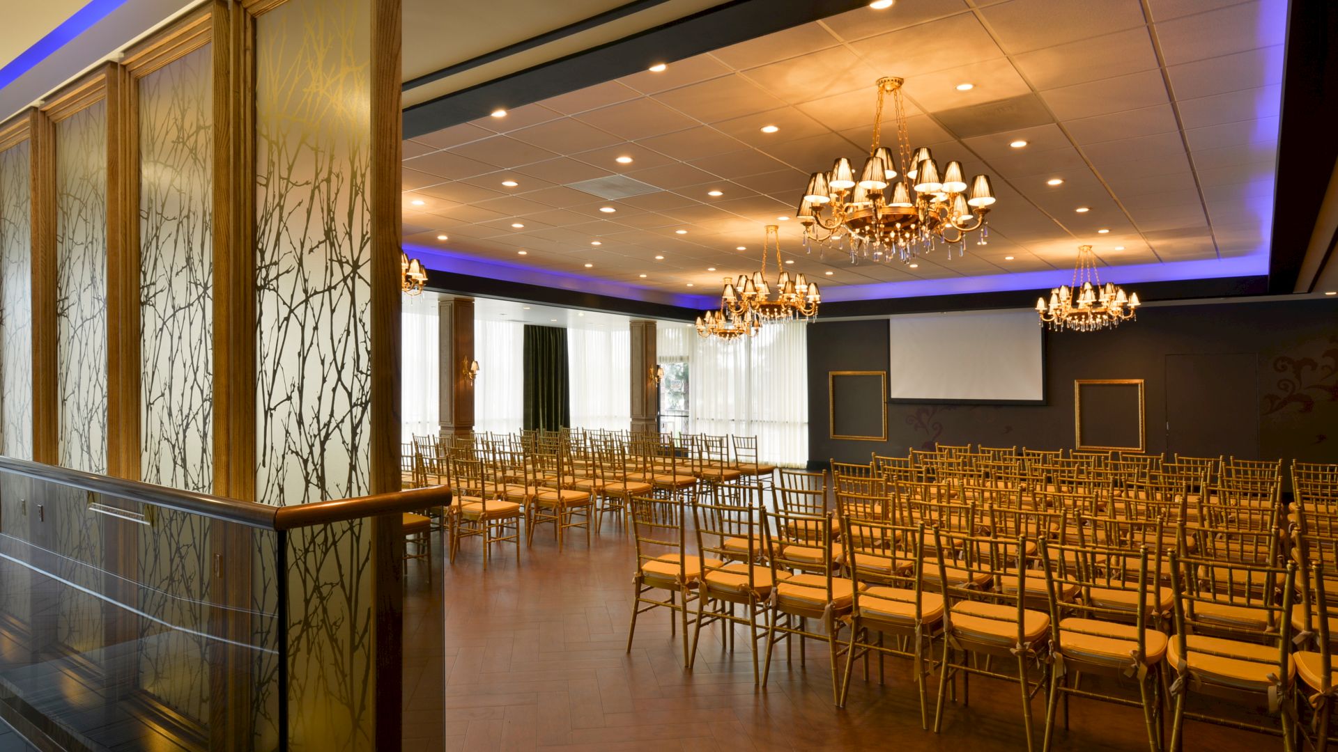 The image shows an elegantly decorated banquet hall with rows of chairs facing a stage area and chandeliers hanging from the ceiling.