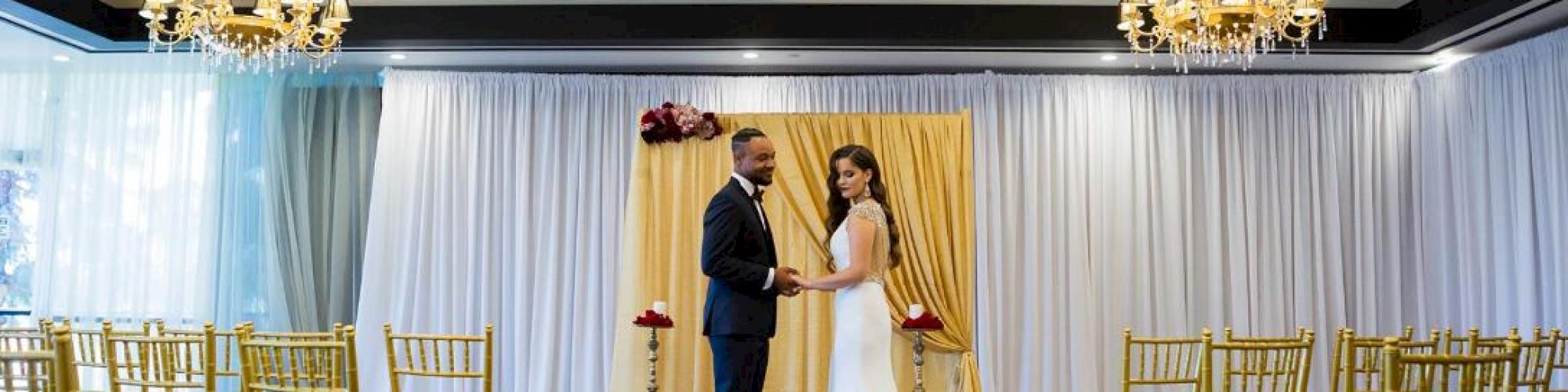 A bride and groom stand at an indoor wedding venue decorated with chandeliers and gold chairs, surrounded by white curtains and candles.