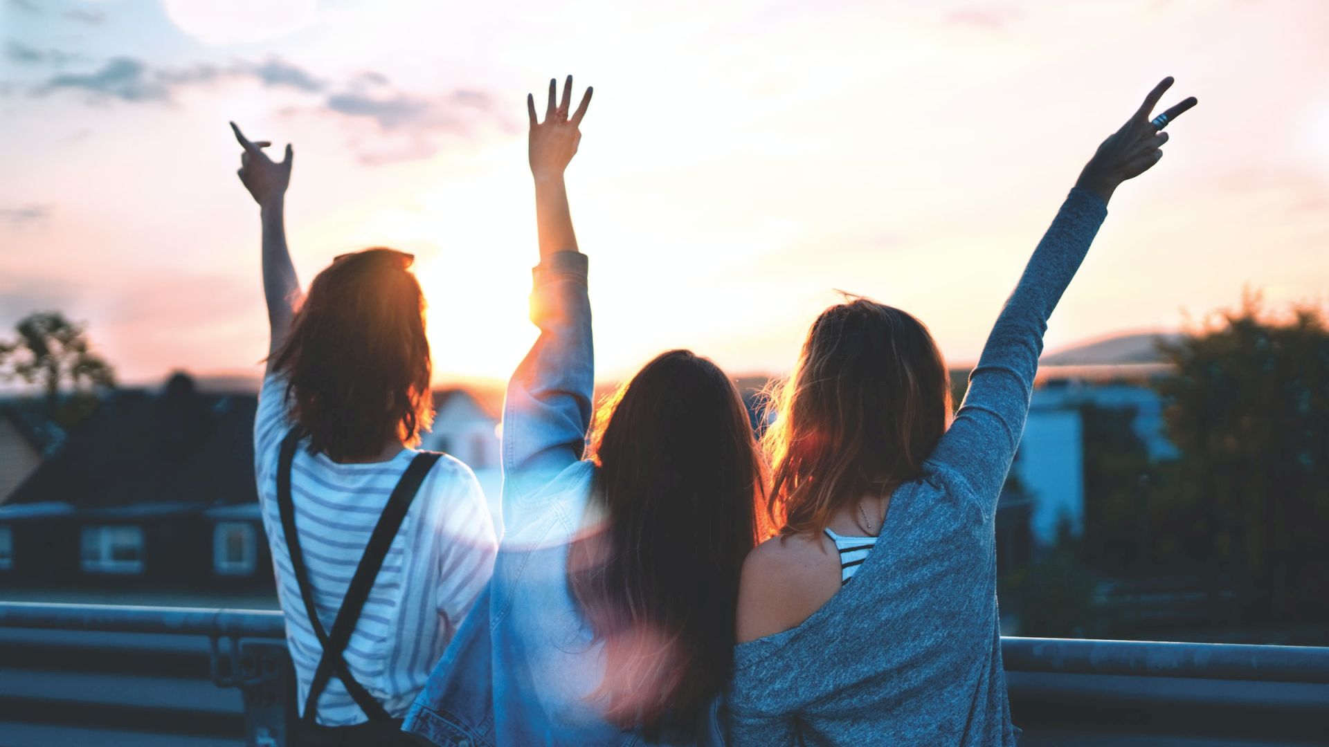 Three people are seen from behind, standing on a rooftop or balcony, facing a sunset. Their arms are raised in celebratory gestures.