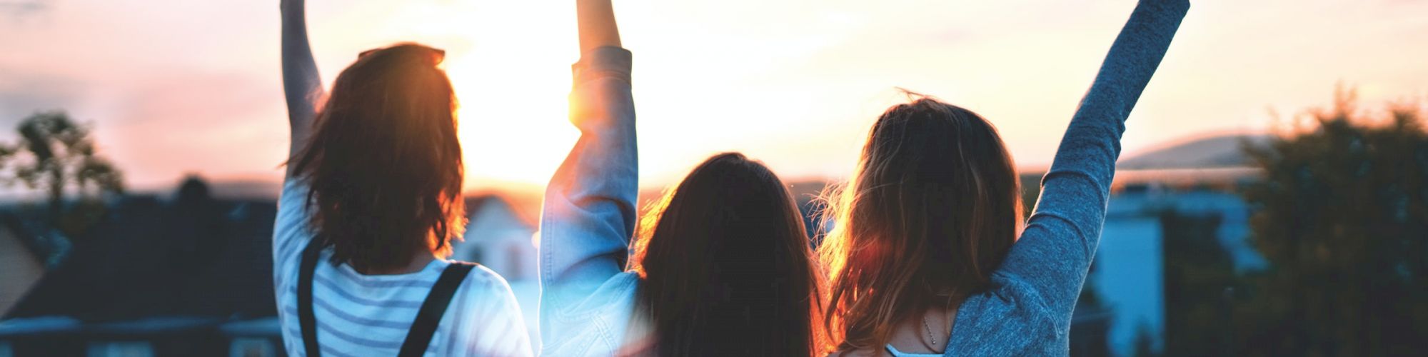 Three people are seen from behind, standing on a rooftop or balcony, facing a sunset. Their arms are raised in celebratory gestures.