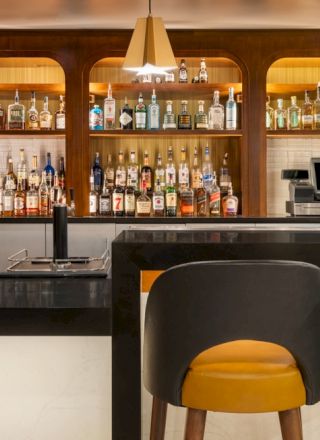 A stylish bar with shelves full of liquor bottles, pendant lights, a marble counter, and two yellow-backed stools.