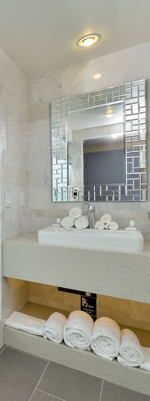 This image shows a neat, modern bathroom with a frosted glass shower, white bathrobe, sink with mirror, and rolled towels on a shelf.