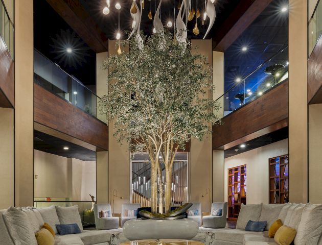 An elegant atrium lounge with a towering indoor olive tree, hanging lights, and modern sofas glowing under a chandelier.