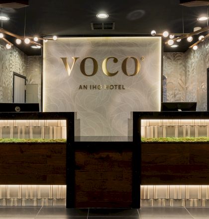 Indoor hotel reception/lobby area with the &ldquo;VOCO&rdquo; and &ldquo;AN IHG HOTEL&rdquo; sign, warm ceiling lights, and decorative bonsai displays.
