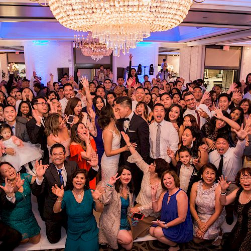 A large group of people, likely at a wedding, are gathered indoors under a chandelier, smiling and waving at the camera.