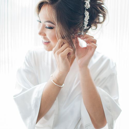 A woman dressed in a white robe is fixing her earring. Her hair is styled with white floral accessories. She has a peaceful expression on her face.