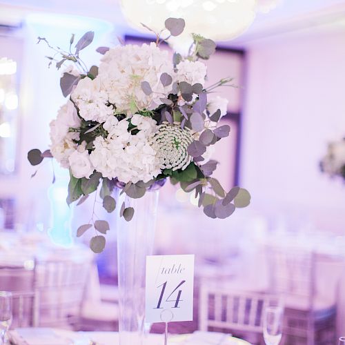 A beautifully decorated table set for an event with a tall floral centerpiece, elegant place settings, and table number 14, under a chandelier.