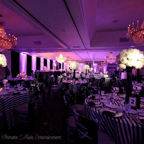 A decorated event hall with purple lighting, chandeliers, and elegantly set tables with floral centerpieces and striped tablecloths.