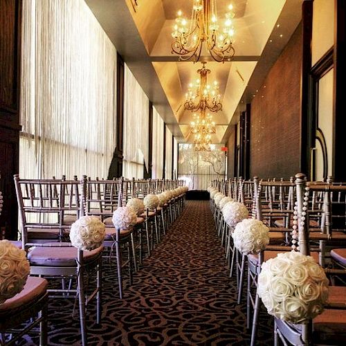 This image shows a wedding ceremony setup with rows of chairs adorned with white flower arrangements, chandeliers, and elegant decor.