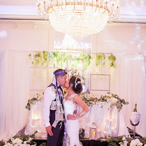 A couple wearing formal attire is standing under a chandelier in a decorated venue, sharing a kiss. The surrounding area is adorned with flowers.