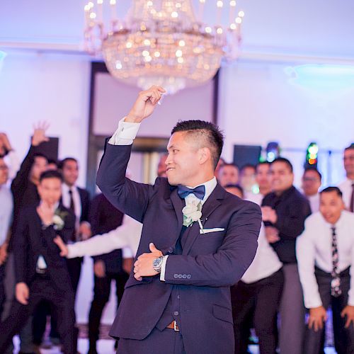 A man in a suit is dancing at an event, with a group of people cheering in the background under a chandelier in a decorated room.