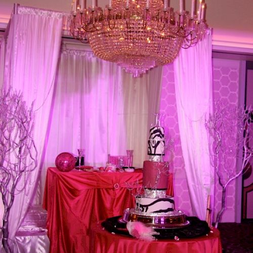 A scene with a chandelier, pink curtains, red tablecloths, and a zebra-striped cake on a table. Decorations include vases and white branches.