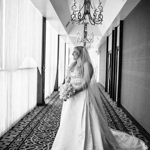 A bride in a white wedding gown stands in a hallway adorned with chandeliers and patterned carpet, holding a bouquet, gazing ahead.