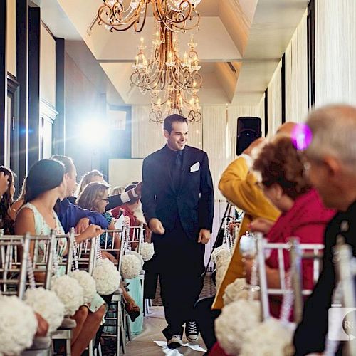 A man walks down an aisle in a well-lit room with chandeliers. Guests are seated on both sides, decorated with white flowers, watching him.