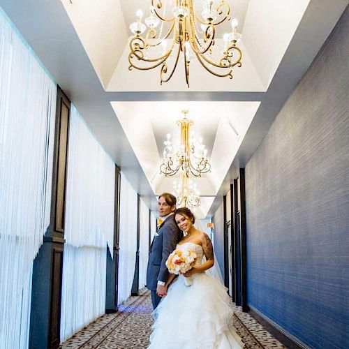 A couple is in a decorated hallway with chandeliers; the woman is in a wedding dress, and the man is in a suit, standing side by side.