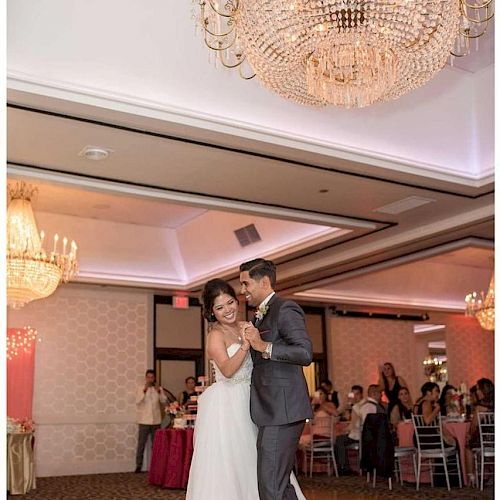 A couple is dancing in a decorated banquet hall with chandeliers. Guests are seated at tables in the background, watching and taking photos.