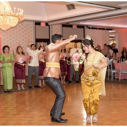 A couple dressed in traditional attire is dancing in a ballroom, surrounded by guests in similar clothing, all under elegant chandeliers, at an event.