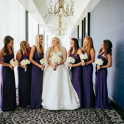 A bride in a white dress stands with five bridesmaids in purple dresses, all holding bouquets, in a decorated hallway with chandeliers.