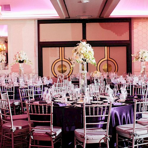 This image shows a fancy banquet hall set up for an event with round tables, elegant centerpieces, and silver chairs under pink lighting.