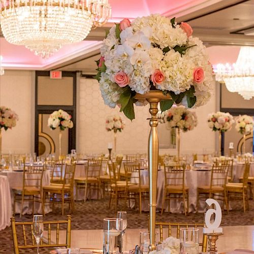 An elegant banquet hall decorated with chandeliers and tall floral centerpieces on tables, set for a formal event with gold-colored chairs.