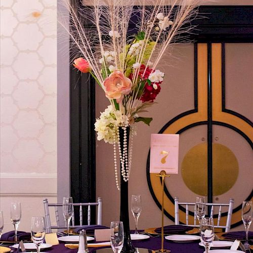 Elegant table setting with a tall floral centerpiece, purple tablecloth, glasses, plates, and silverware in a chic, decorated venue.