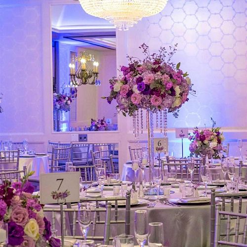 An elegant banquet hall is decorated with flower centerpieces, round tables set with plates and glasses, and a large chandelier hanging from the ceiling.