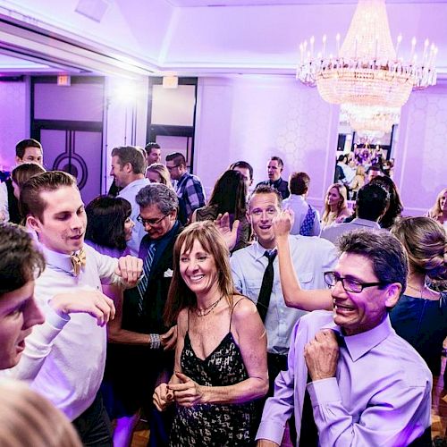 People are dancing and socializing at a lively indoor event with purple lighting and a chandelier, creating a festive atmosphere.