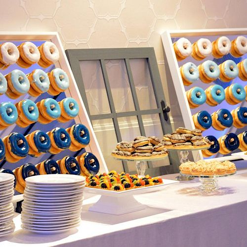 A dessert table with assorted doughnuts on display racks, other pastries, and stacks of plates. The background features a decorative window pane.