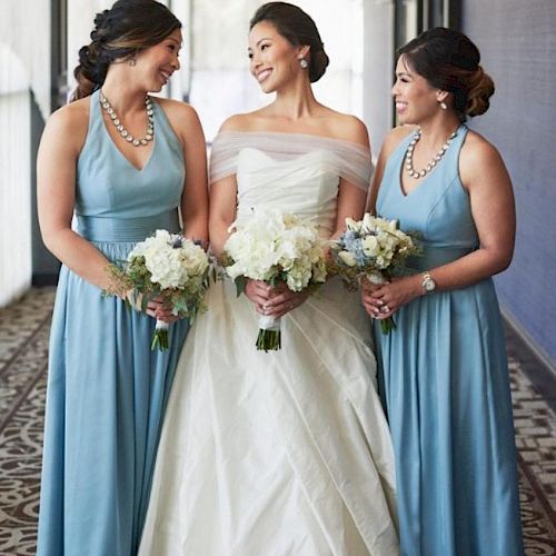 A bride in a white gown stands between two bridesmaids in blue dresses, all holding bouquets and smiling at each other in a brightly lit hallway.