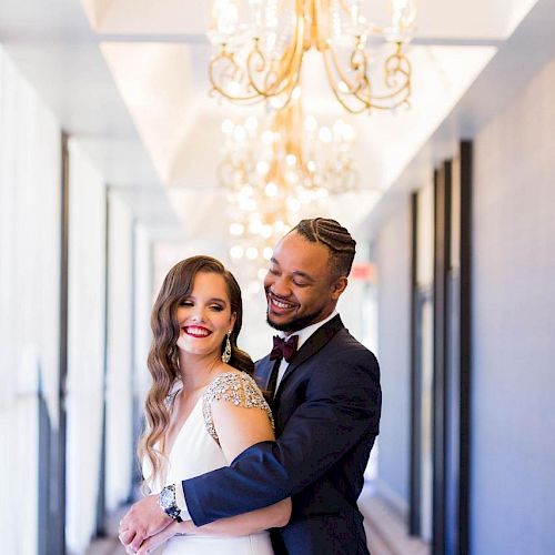 A smiling couple in wedding attire stands in an ornate hallway with chandeliers, the groom embracing the bride from behind.