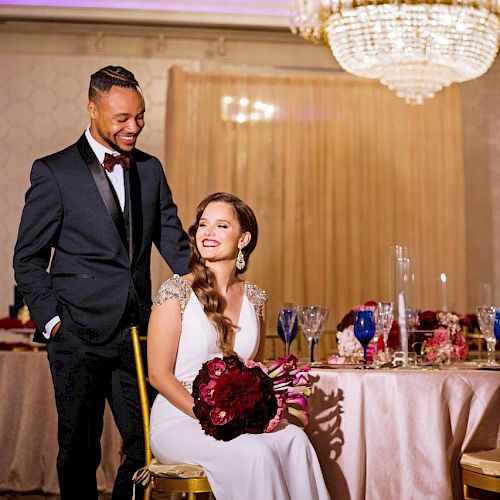 A couple dressed formally, with the woman seated holding a bouquet, under an elegant chandelier in a decorated dining setting.