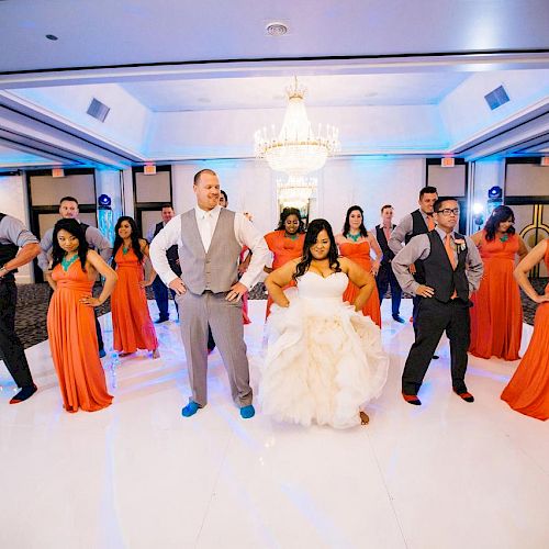 A wedding party is dancing in a ballroom, with the couple in the center. The bridesmaids wear orange dresses, and the groomsmen wear gray vests.