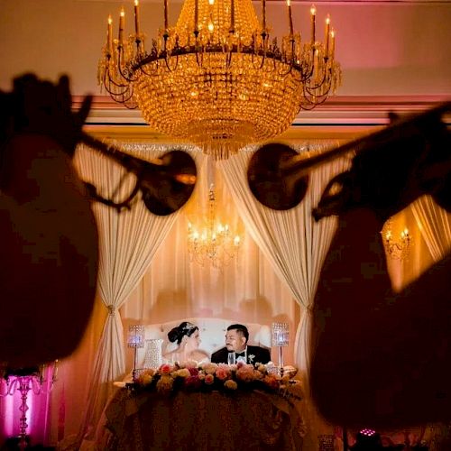 A couple is seated at a decorated table under a chandelier while musicians play trumpets in the foreground, creating a romantic atmosphere.