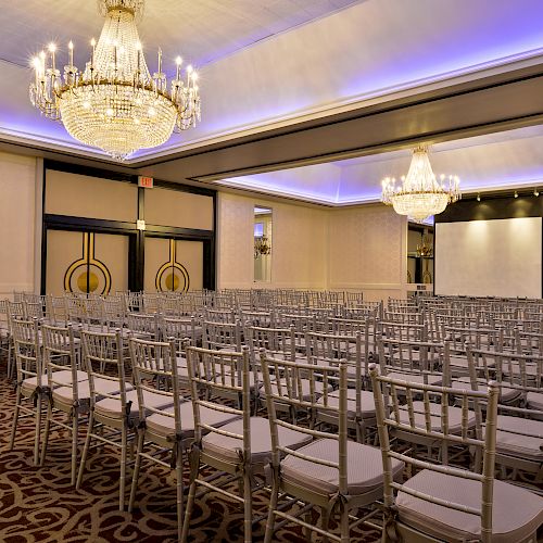 An elegant conference room with rows of chairs facing a projection screen, featuring chandeliers and decorative lighting on the ceiling.