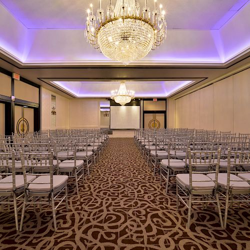 A spacious, elegant ballroom setup with rows of chairs facing a projection screen, adorned with chandeliers and purple ceiling lighting.